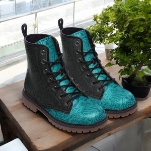 A pair of black and teal patterned boots are displayed on a wooden surface, with a potted plant and a window in the background.