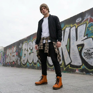 A young man wearing a black jacket, plaid shirt, and orange boots stands in front of a graffiti-covered wall.