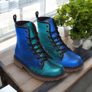 A pair of blue and teal-colored boots with black laces, placed on a wooden surface near a window with greenery outside.