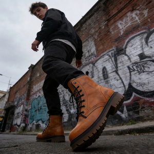A person wearing black pants and brown boots stands in front of a graffiti-covered brick wall.