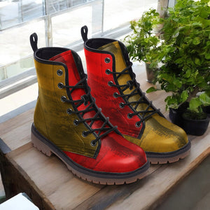 A pair of colorful, patterned boots with red and yellow hues are displayed on a wooden surface, with a potted plant in the background.
