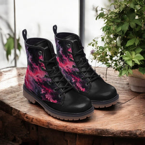 A pair of black boots with a vibrant, abstract pattern in shades of pink and purple, displayed on a wooden surface next to a potted plant.