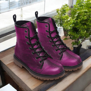 A pair of purple combat boots with black laces and soles, placed on a wooden surface near a window with green plants.